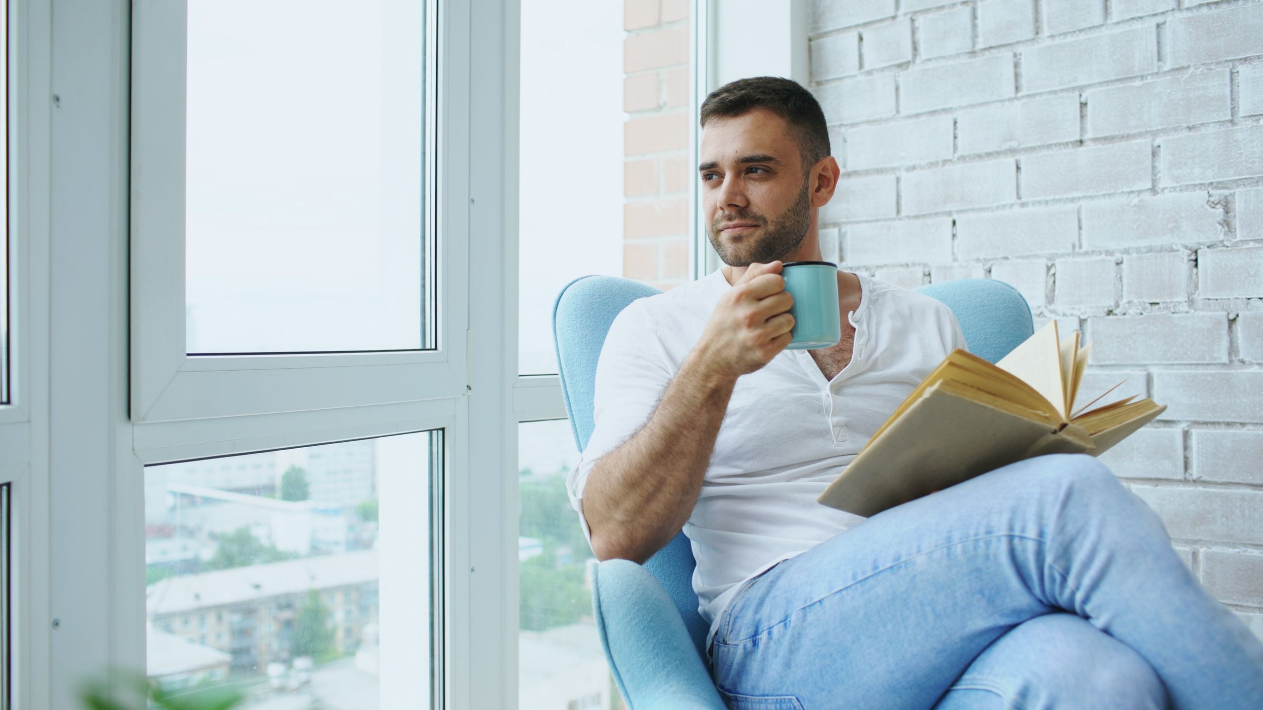 Man enjoys a quiet moment with coffee and a book in a cozy chair by the window.