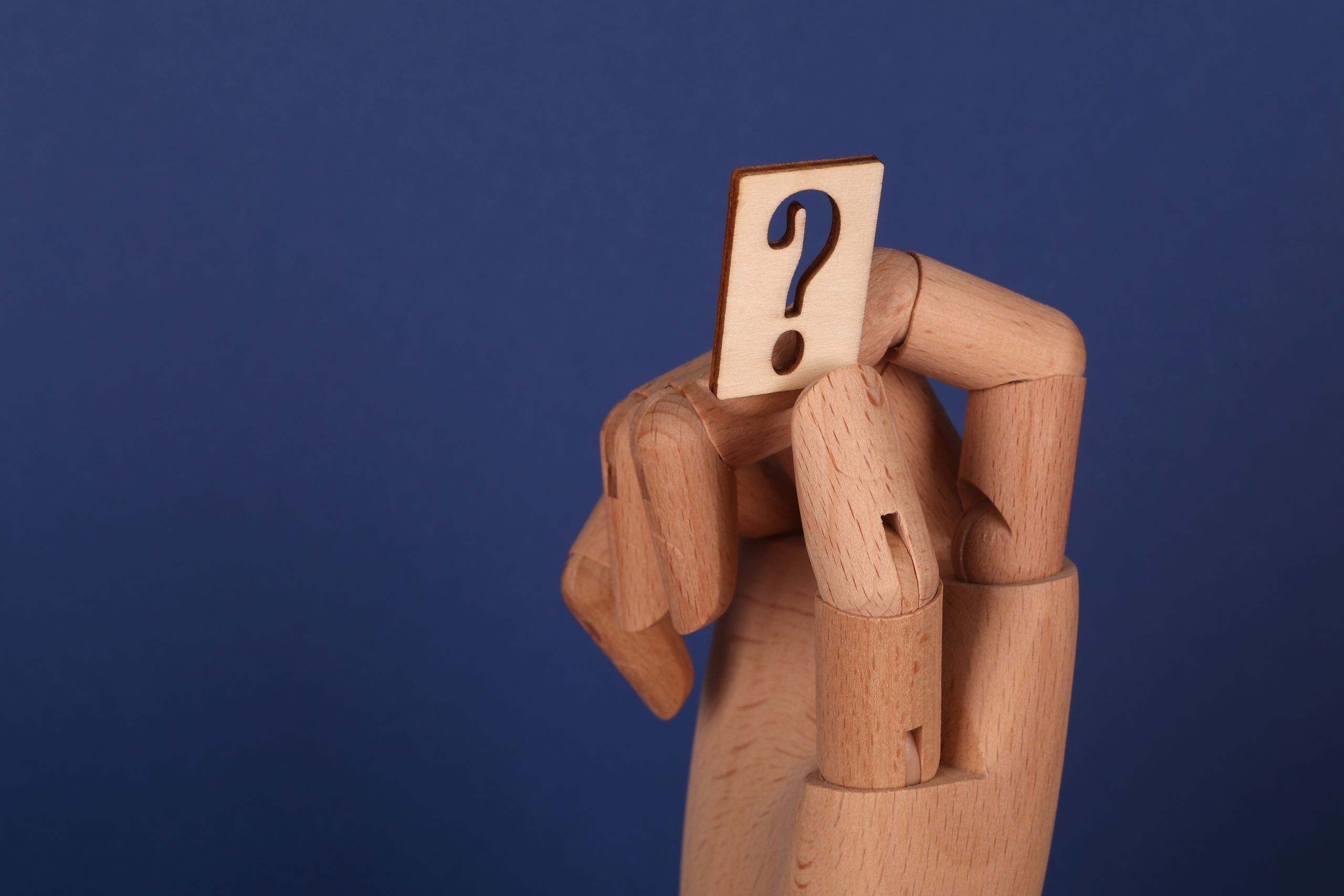 A wooden hand model holding a cut-out question mark against a blue background, symbolizing curiosity or inquiry.