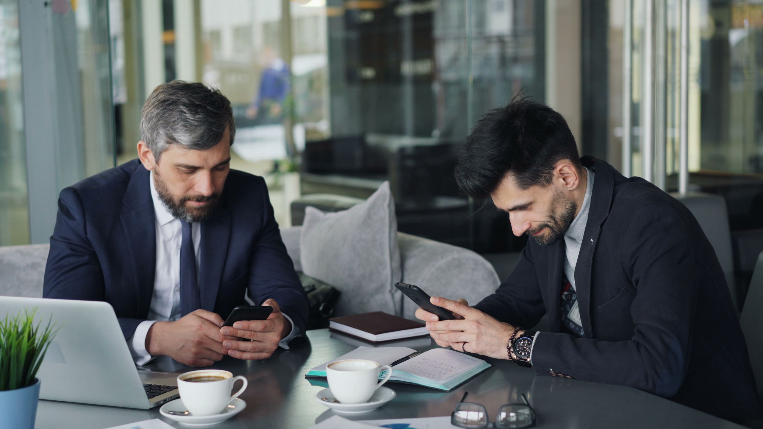 Two businessmen in suits using smartphones at a café table with laptops and coffee.